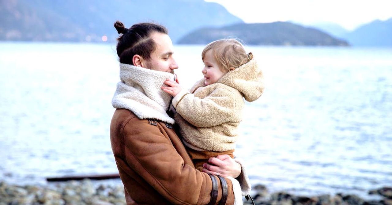 Side view of happy father and little kid in warm outwear on hands standing on stony coast near water and looking at each other during vacation