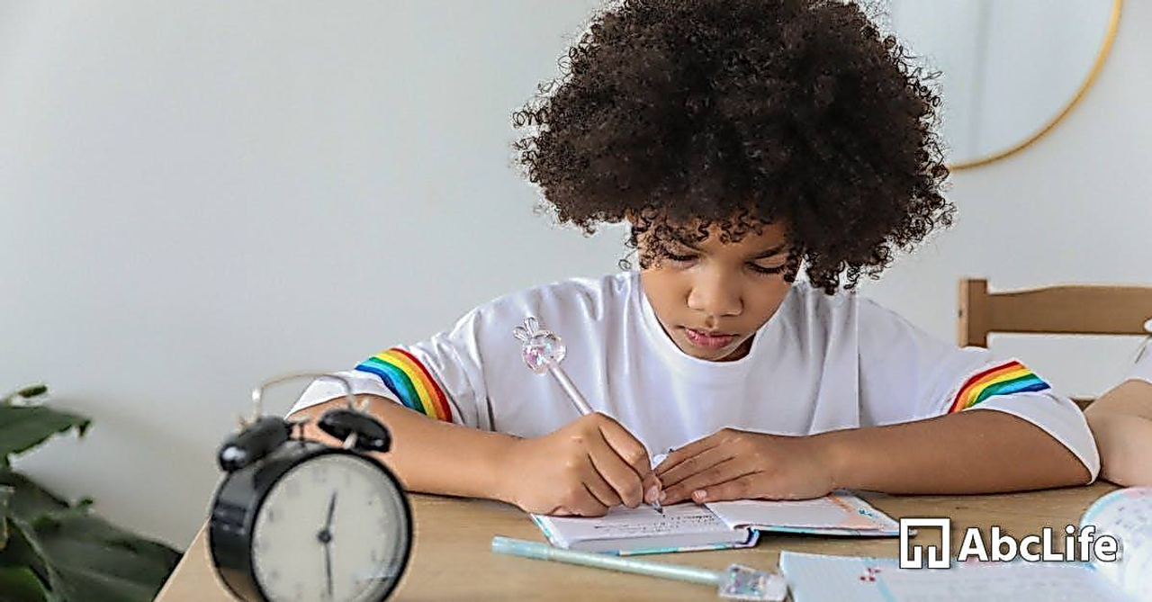 Concentrated African American child writing in notebook while studying at desk with alarm clock at home