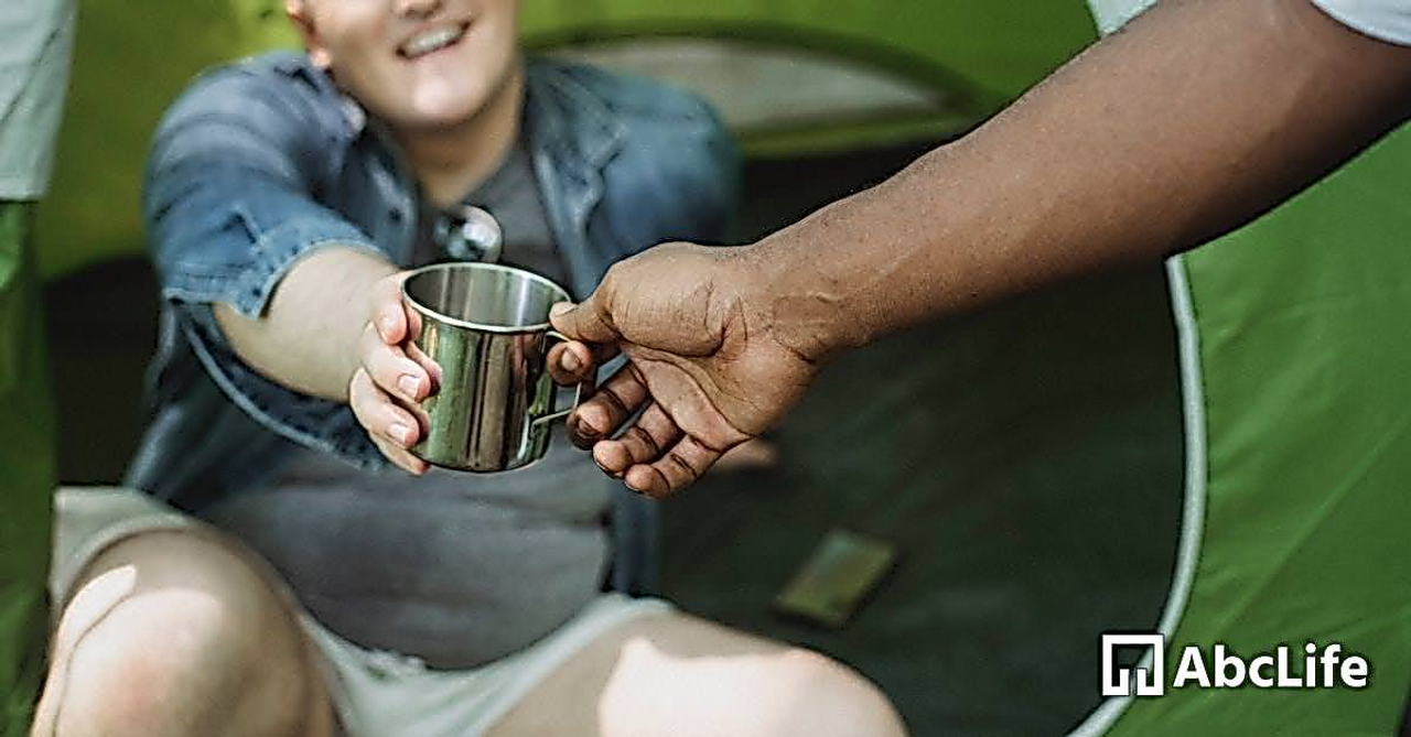 Crop anonymous African American man taking camping metal cup from cheerful friend sitting in tent in daytime