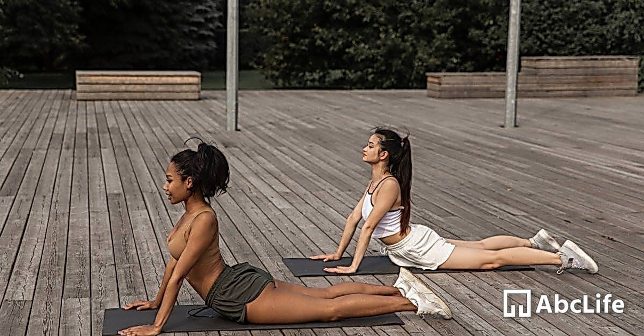 Side view of focused diverse girlfriends doing High Cobra pose while stretching bodies on wooden platform in park