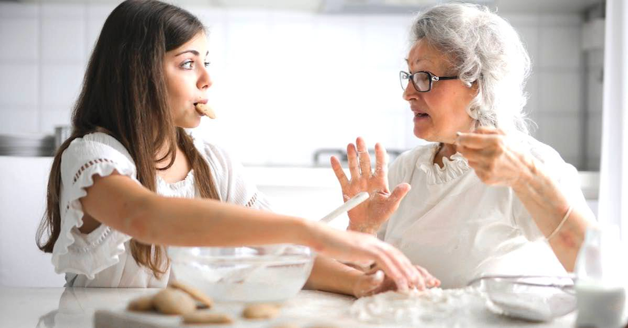 Calm senior woman and teenage girl in casual clothes looking at each other and talking while eating cookies and cooking pastry in contemporary kitchen at home