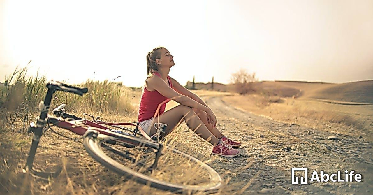 Full body of female in shorts and top sitting on roadside in rural field with bicycle near and enjoying fresh air with eyes closed
